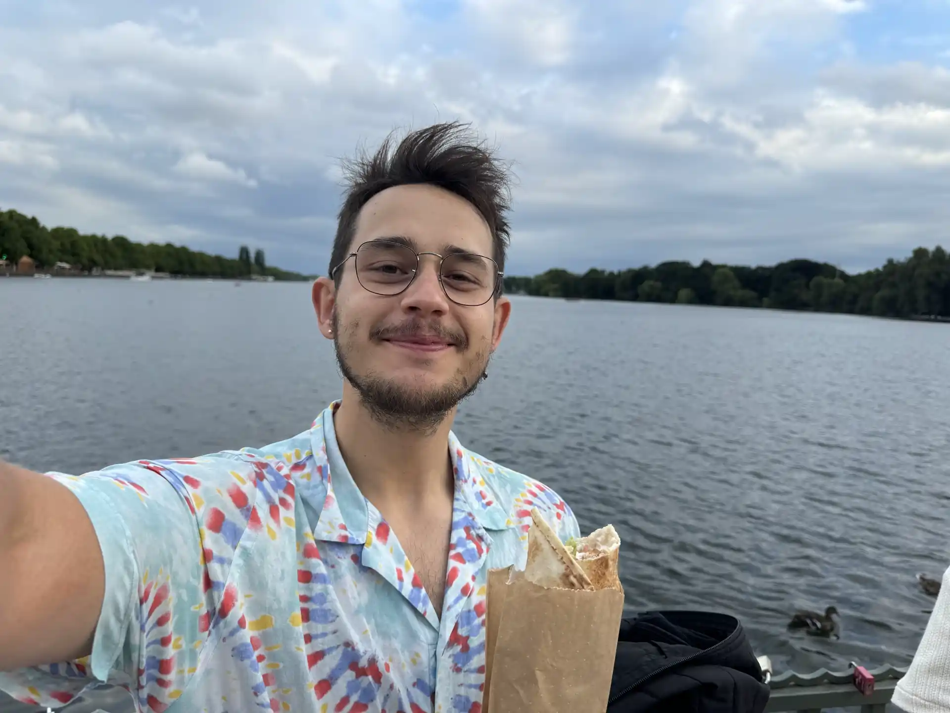 In the middle of the picture, René Bergenroth, who is slightly smiling, is wearing glasses and is a bit rounder with earrings and a beard. He is wearing a shirt in various colors, with a cool design featuring a lot of red, yellow, and blue dots. He is holding a wrap, and in the background, you can see the horizon at the Marschseefest. The sky is cloudy, but it's a warm day.