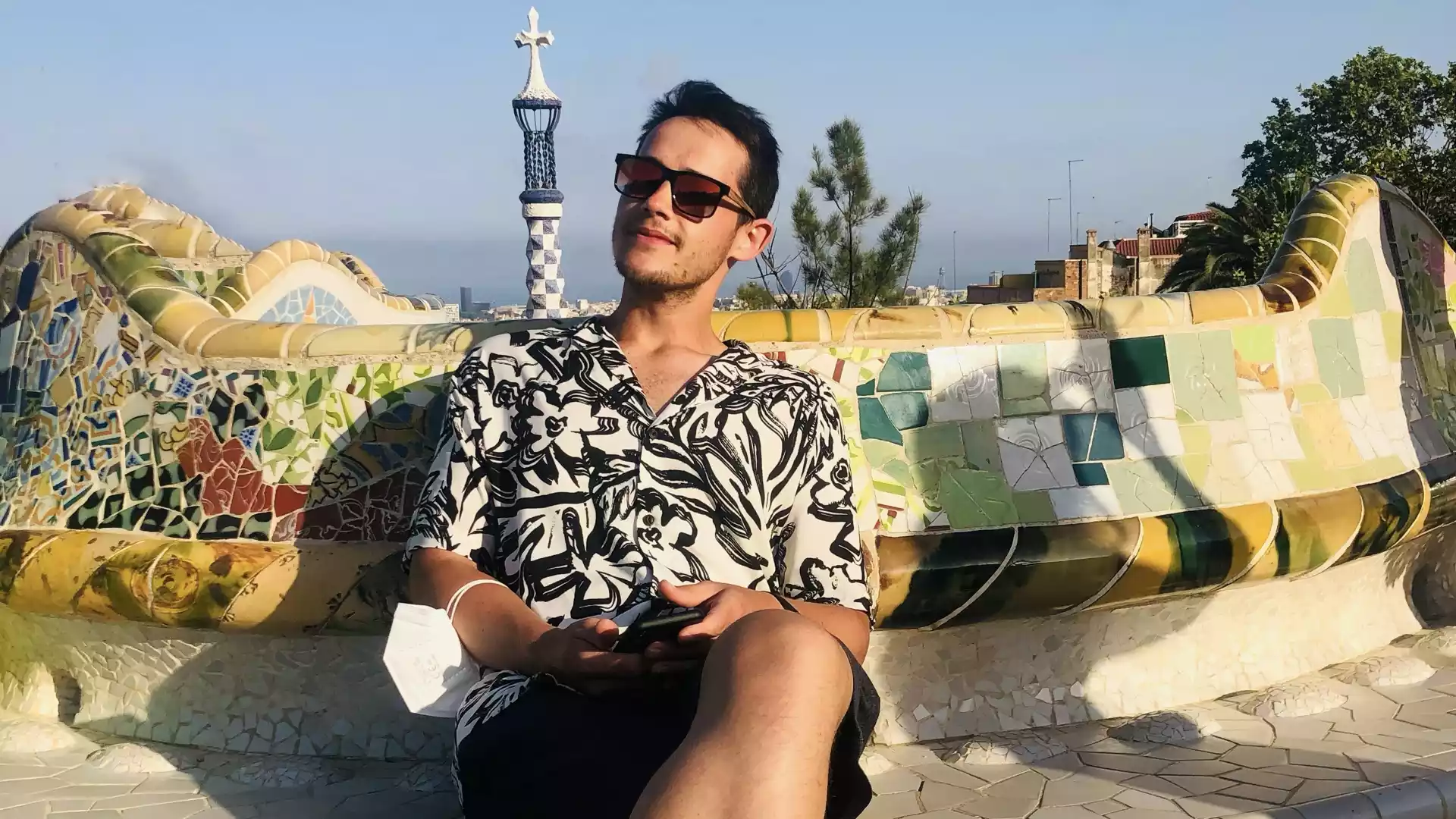 A man René Bergenroth with a three-day stubble, wearing sunglasses and a white-and-black patterned shirt, paired with black shorts. He is sitting on a mosaic bench in a park in Barcelona, enjoying the warm weather at sunset. René Bergenroth