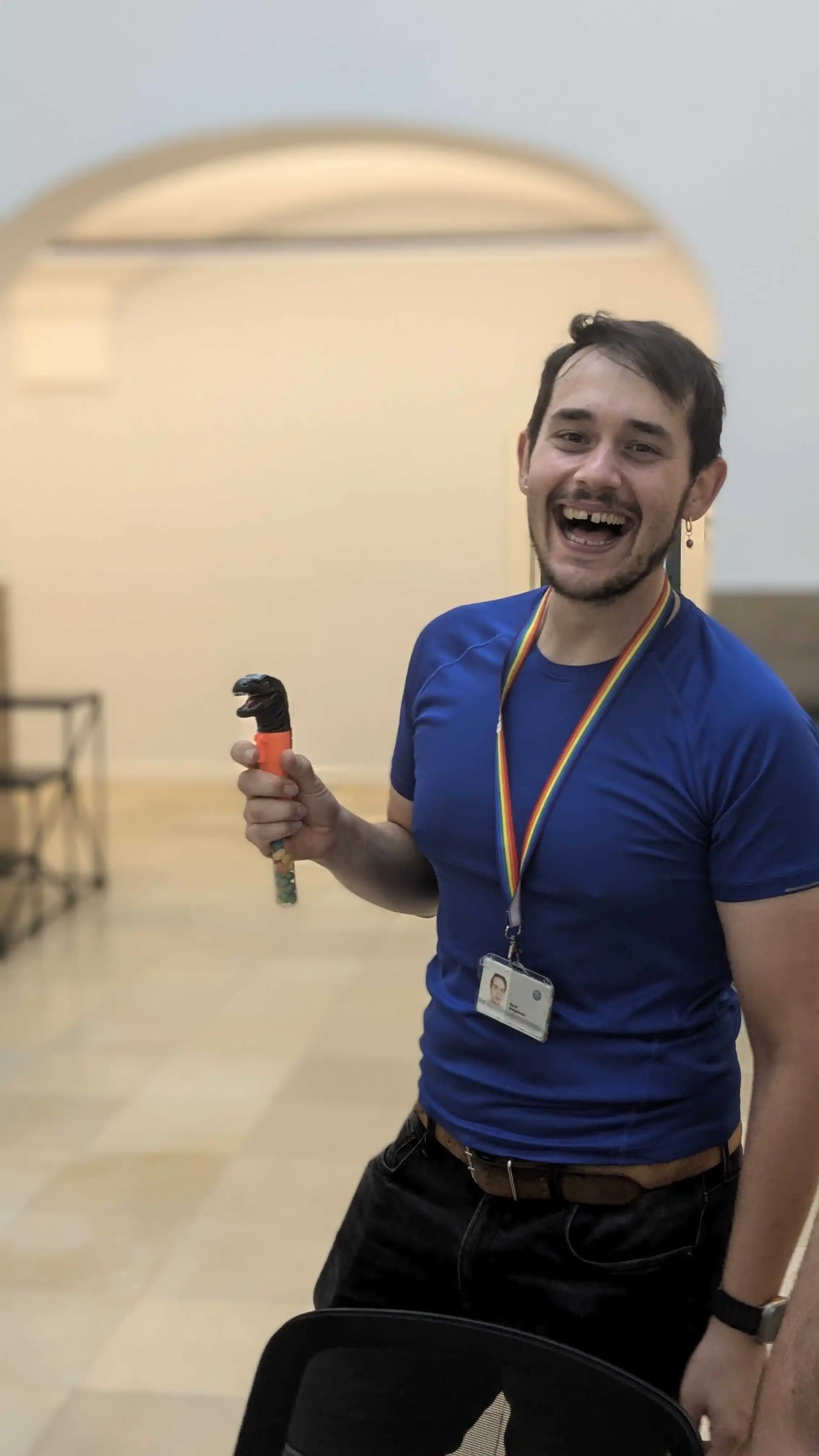 René Bergenroth holding a toy, In der Hanumag building, Der Hintergrund ist ziemlich blöd. He is wearing a skinny blue training shirt. He looks really happy and he is smiling. His hair is not so in a good shape because he is sweating. He wearing black trousers. The toy is a small dinosaur with a handle and it's only the head like a Smaller stick. Und Sie können am Ende sehen, die Zucker-Candies in dieser Kiste.